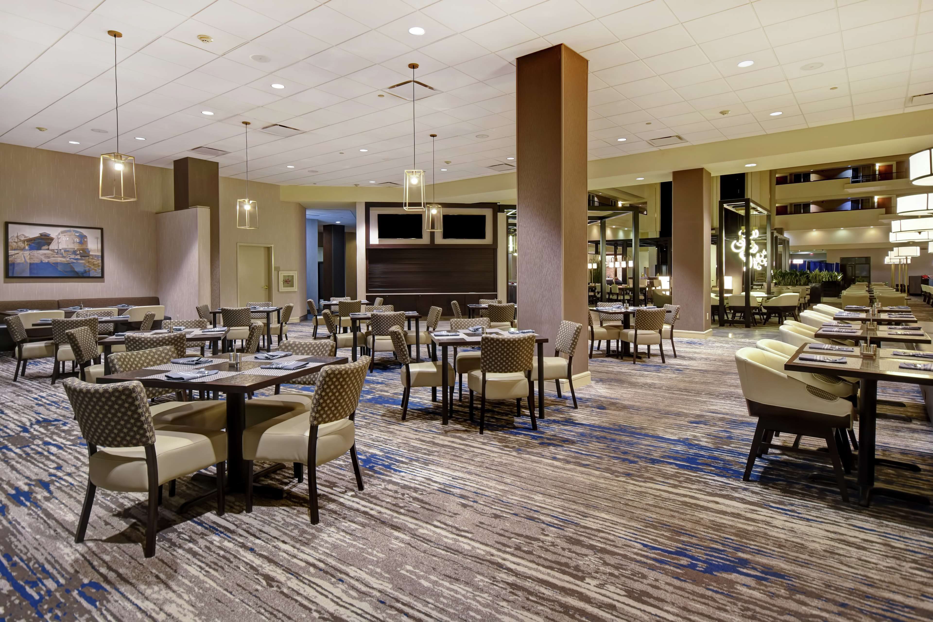 A hotel dining area with multiple tables and chairs, a large screen TV, and a patterned carpet.