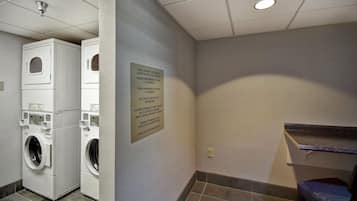 Un espace buanderie équipé d’une pile de lave-linge et de sèche-linge, un panneau indiquant les règles d’utilisation de la buanderie pour les clients, et un banc.
