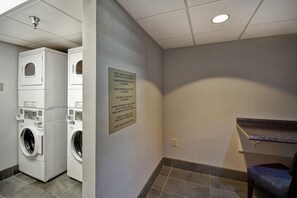 A laundry area with stacked washers and dryers, a sign detailing guest laundry policies, and a bench. - Embassy Suites by Hilton Montgomery Hotel & Conference Ctr (Montgomery)