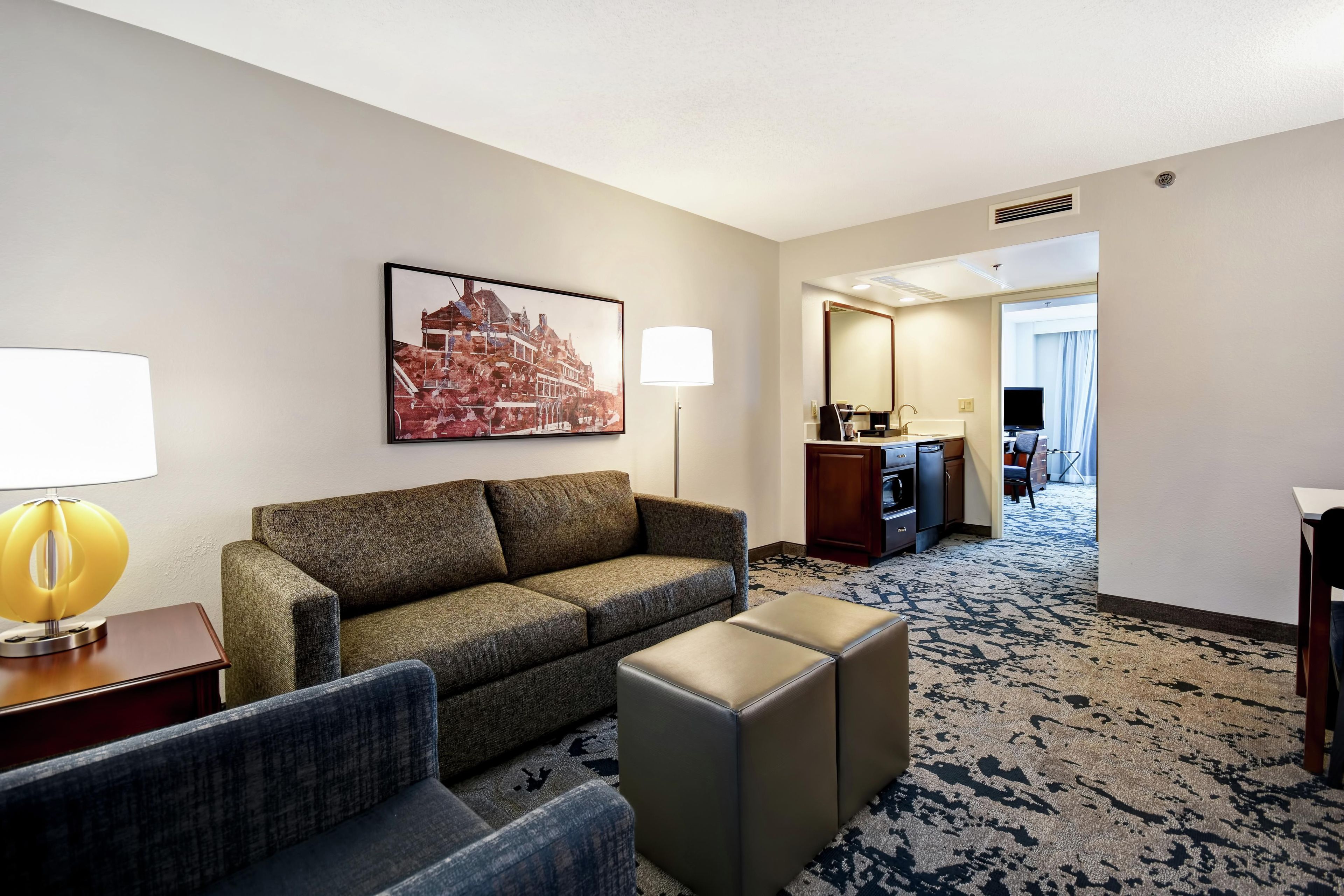 A hotel room with a sofa, ottoman, lamp, and a painting on the wall.