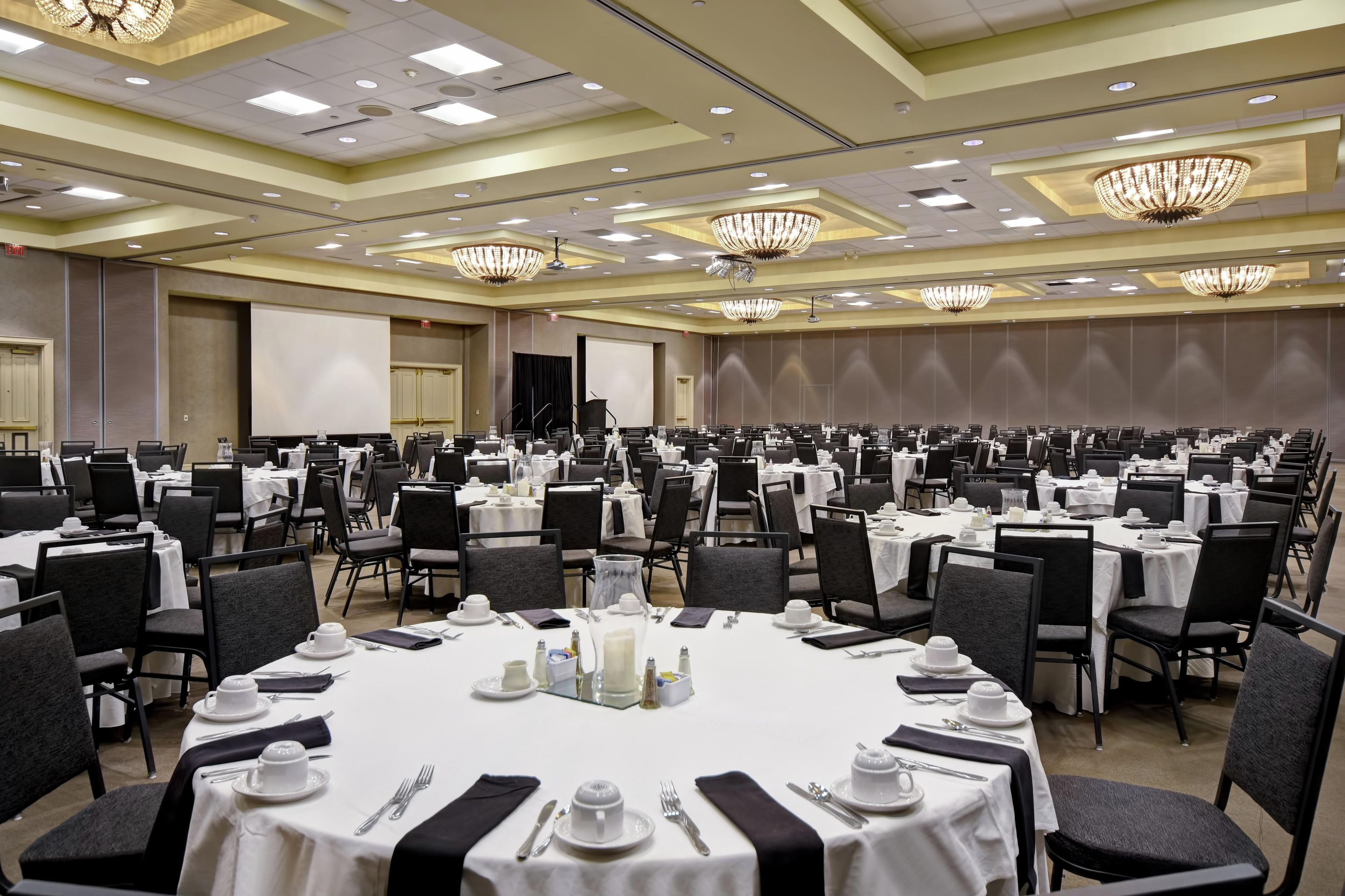 A large conference room with multiple round tables, each set with white tablecloths, chairs, and silverware.