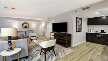 Una sala de estar moderna con sofá, sillones, una mesa de centro, televisor y una zona de cocina con armarios y electrodomésticos.