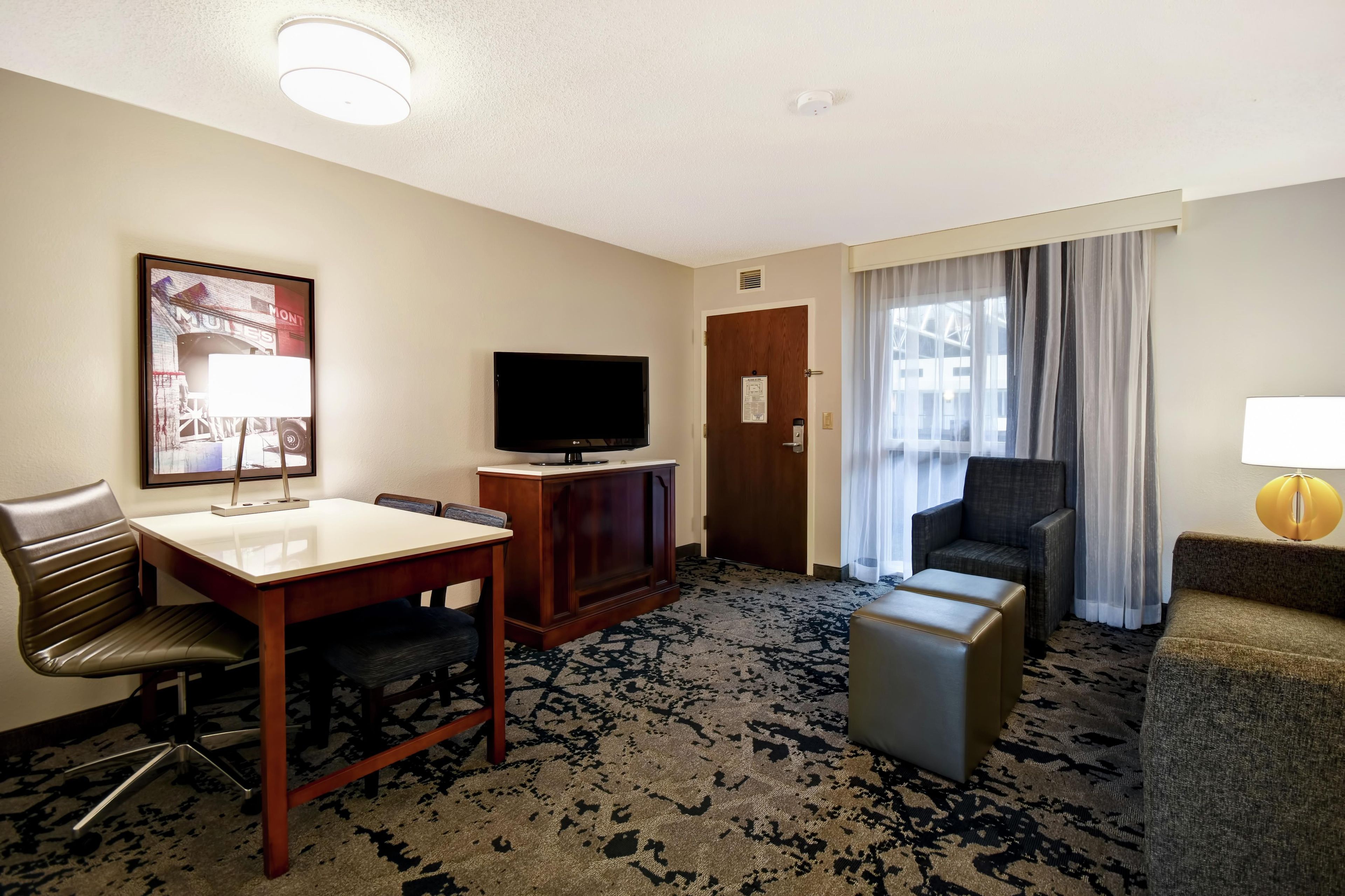 Une chambre d’hôtel équipée d’un téléviseur à écran plat, d’un bureau en bois, d’un fauteuil en cuir, d’un canapé, d’un petit pouf et d’une grande fenêtre avec des rideaux.