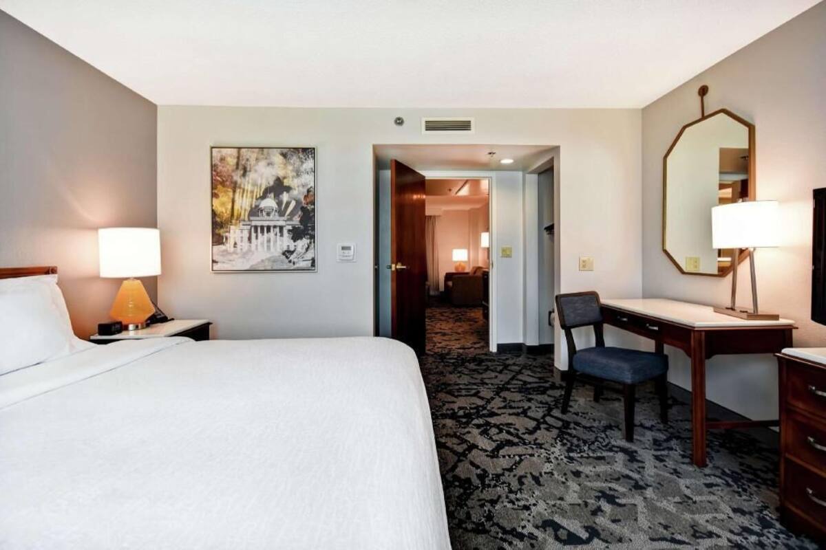 A hotel room with a bed, desk, chair, and a large mirror. There is a painting on the wall and a carpeted floor.