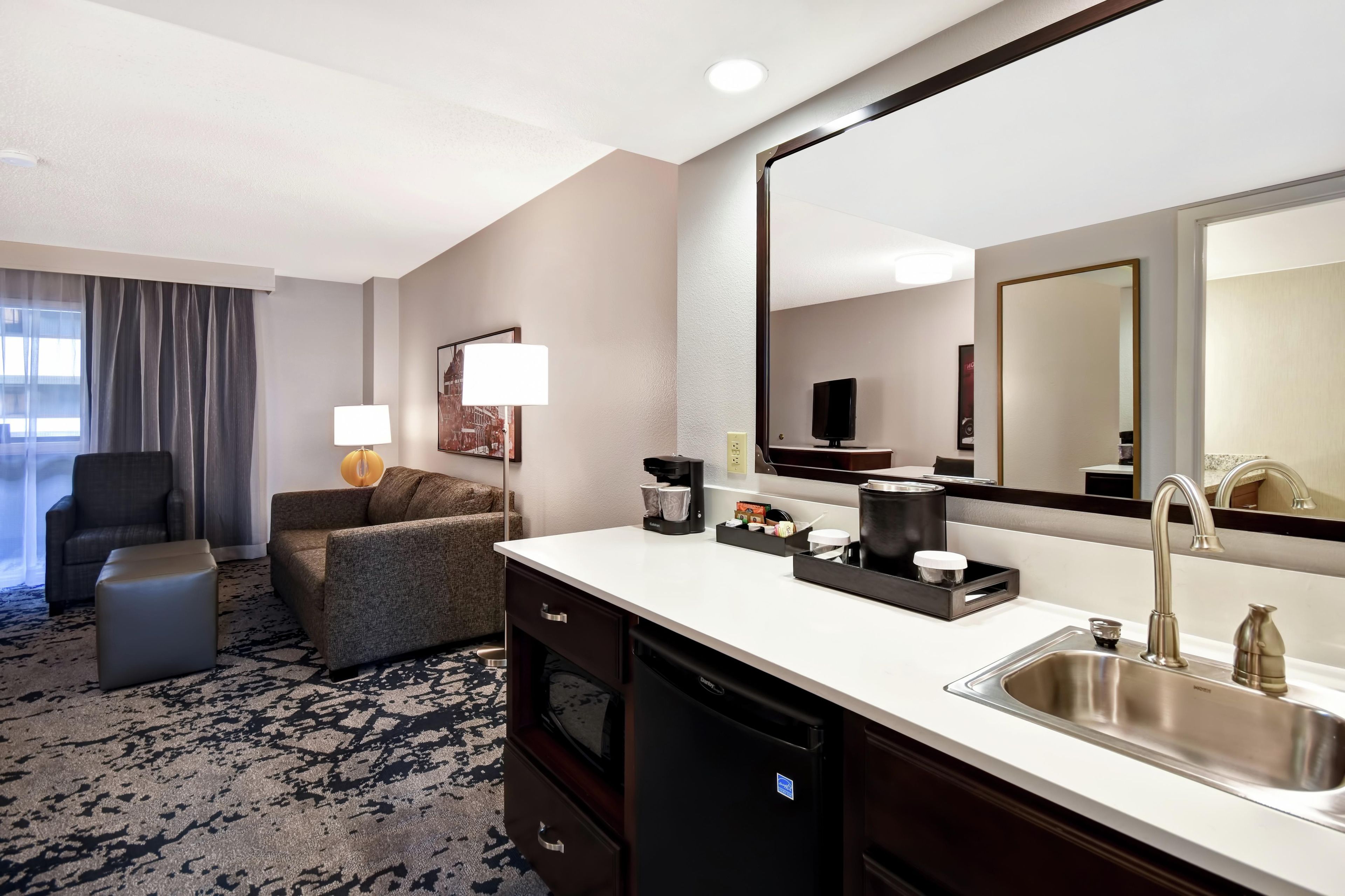 A hotel room with a sofa, a chair, a desk with a sink, a mirror, and a window with curtains.