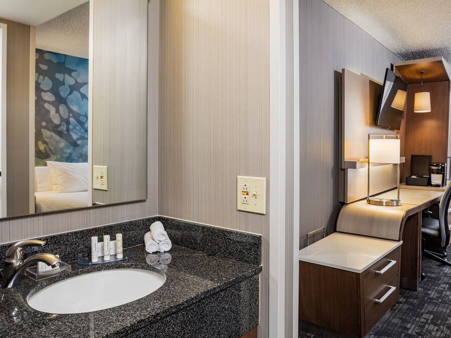 Un baño de hotel moderno con lavamanos de granito, un espejo, un televisor de pantalla plana y un escritorio con una silla.