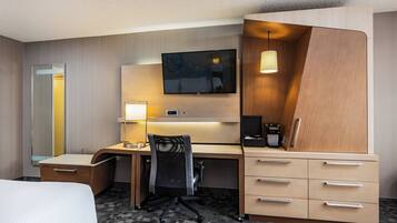 A hotel room with a bed, a desk with a chair, a TV mounted on the wall, a lamp, and a minibar.