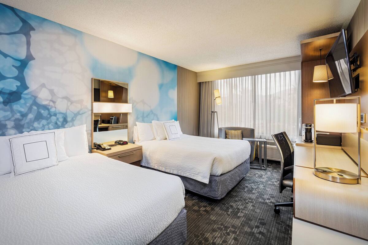 A hotel room with two beds, a desk, a chair, a TV, and a large window with curtains.