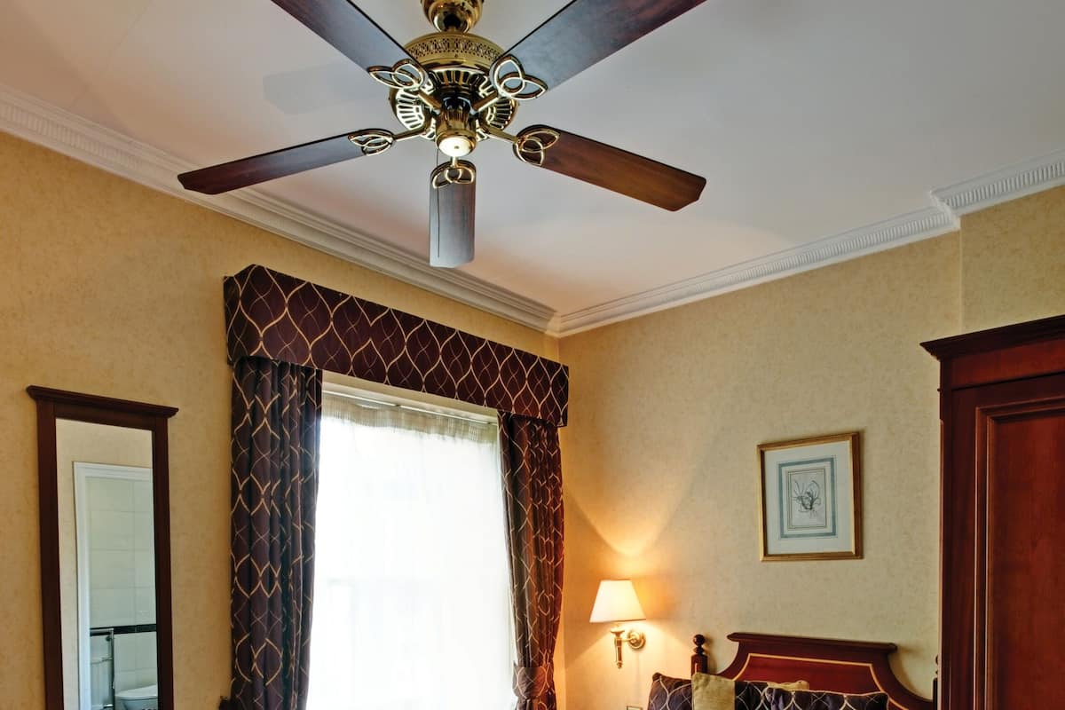 Ein Hotelzimmer mit Bett, Deckenventilator, Fenster mit Vorhängen, Nachttisch mit Lampe und Spiegel.