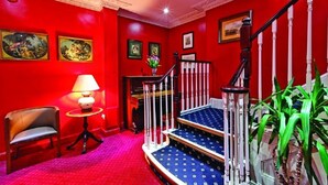 A staircase with a red wall, a wooden chair, a small table with a lamp, and framed pictures on the wall. - Grange Blooms Hotel (London)