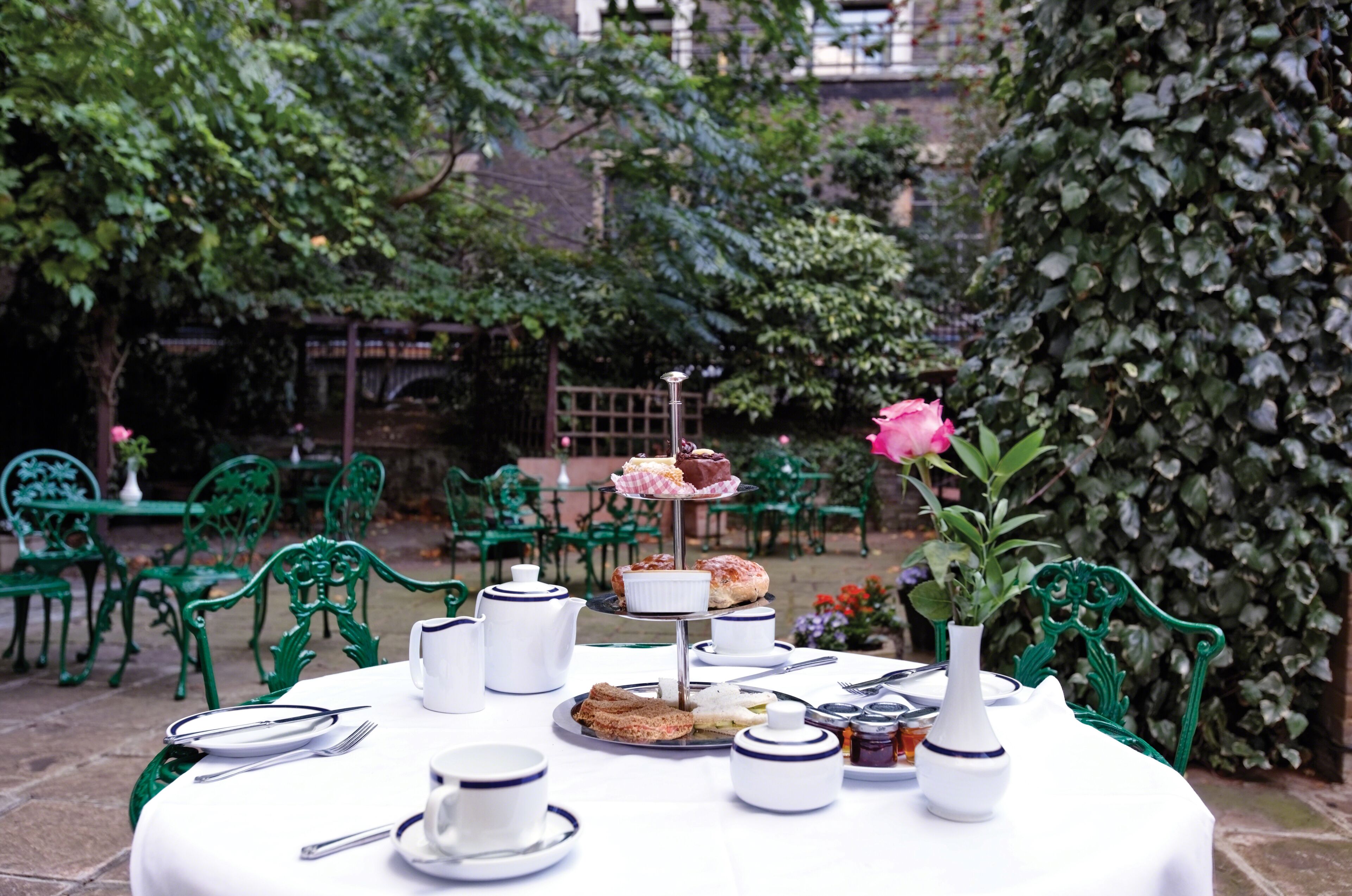 A round table set with tea service, sandwiches, and a rose in a vase, surrounded by green chairs in an outdoor garden.