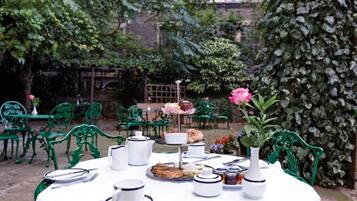 A round table set with tea service, sandwiches, and a rose in a vase, surrounded by green chairs in an outdoor garden.