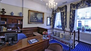 A room with a desk, computer, bookshelf, framed painting, and a view of the outside through large windows. - Grange Blooms Hotel (London)