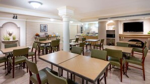 A hotel dining area with multiple tables and chairs, a television mounted on the wall, and a fireplace. - La Quinta East Deerfield Beach - Boca Raton (Deerfield Beach)