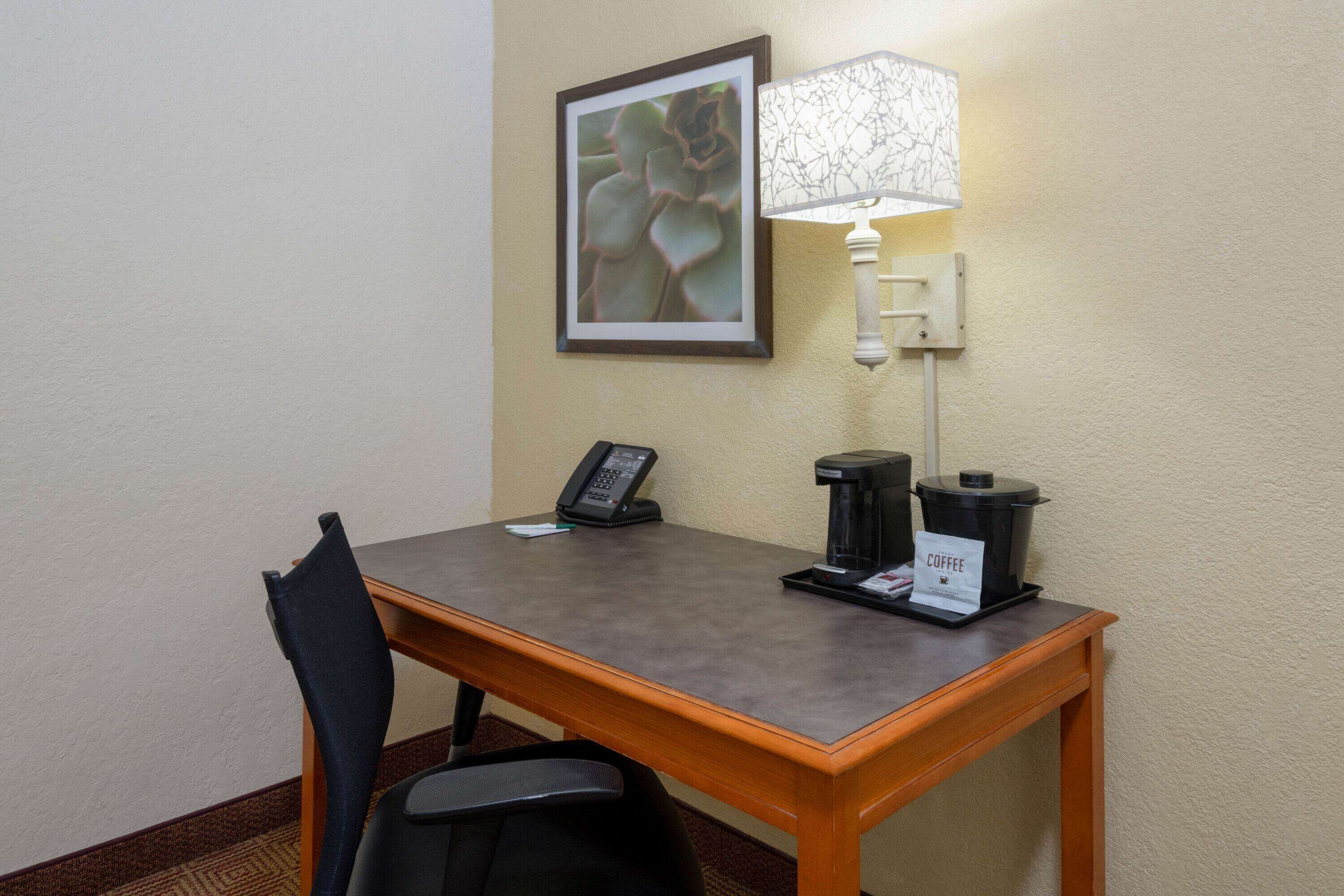A hotel room with a wooden desk, a telephone, a coffee maker, and a framed picture on the wall.