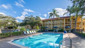 A swimming pool with lounge chairs and umbrellas, surrounded by a fence and palm trees. - La Quinta East Deerfield Beach - Boca Raton (Deerfield Beach)