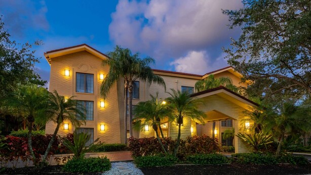 A two-story building with a beige facade, multiple windows, and a red roof. There are palm trees and other greenery in front. - La Quinta East Deerfield Beach - Boca Raton (Deerfield Beach)