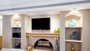 A modern living room with a flat-screen TV mounted above a fireplace, wooden shelves with decorative items, and a microwave in a cabinet. - La Quinta East Deerfield Beach - Boca Raton (Deerfield Beach)