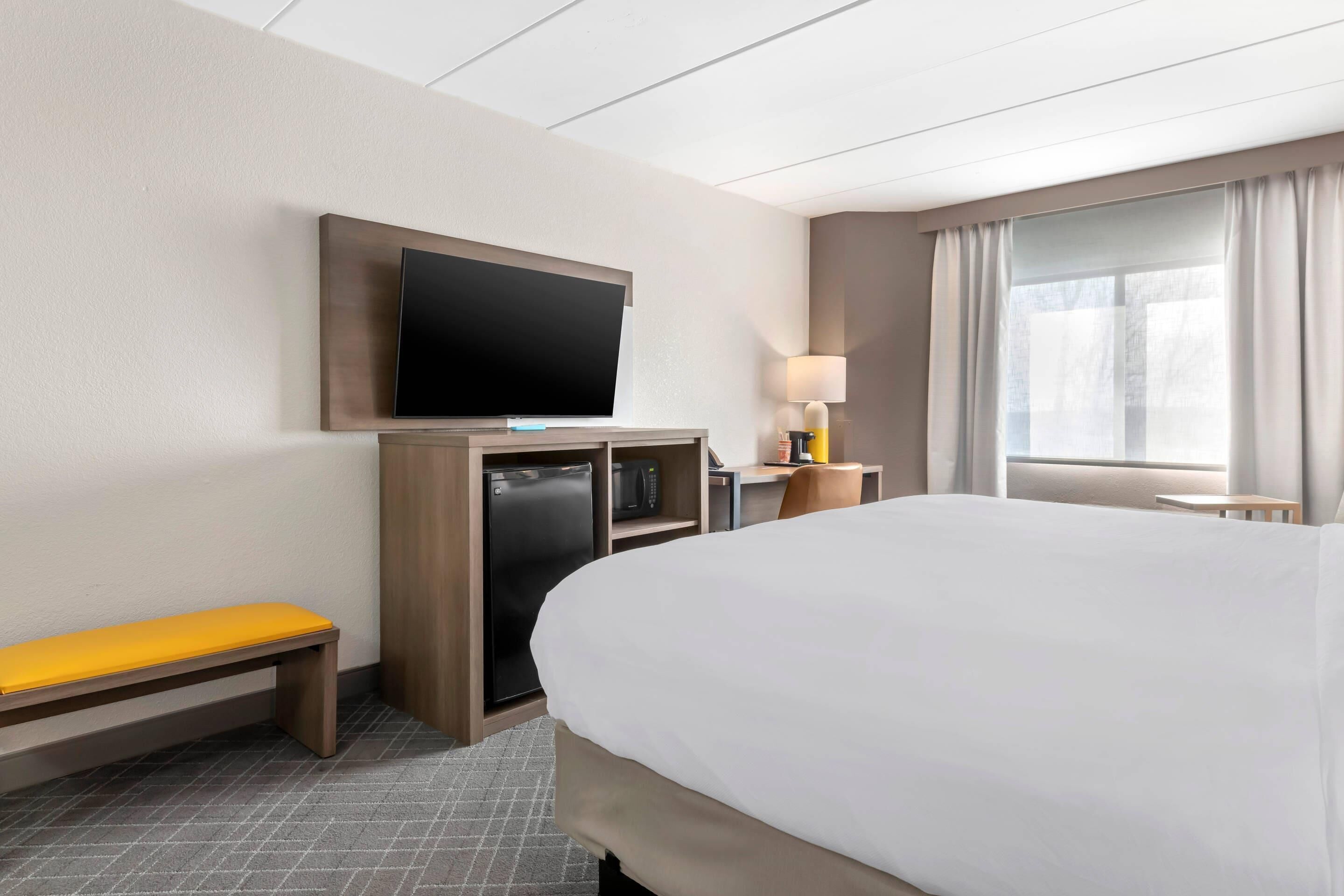 Ein Hotelzimmer mit Bett, flachbildfernseher an der Wand, Schreibtisch mit Lampe und Sitzbank.