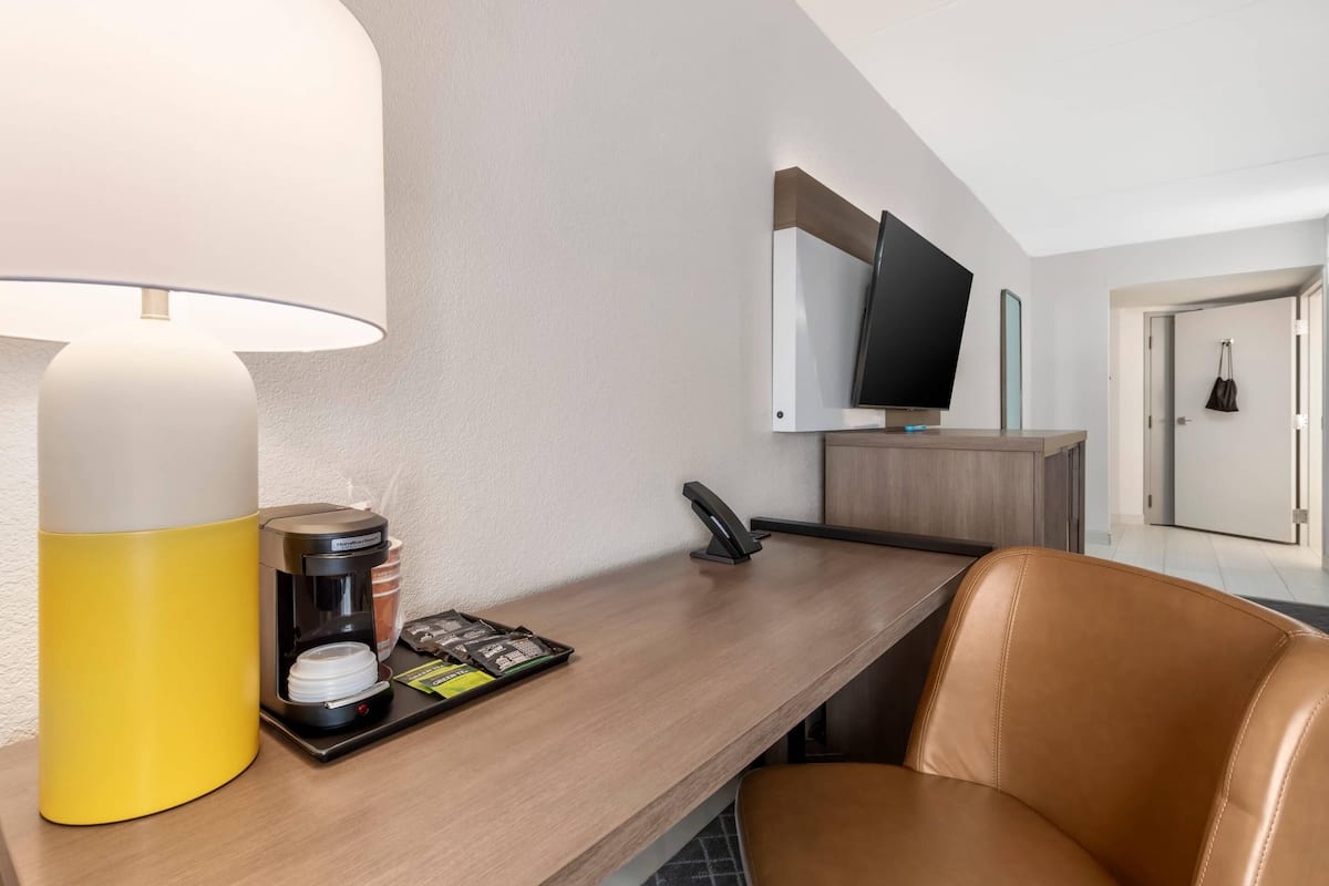 A hotel room with a desk, a lamp, a coffee maker, a tray with coffee pods, a phone, and a television on a stand.