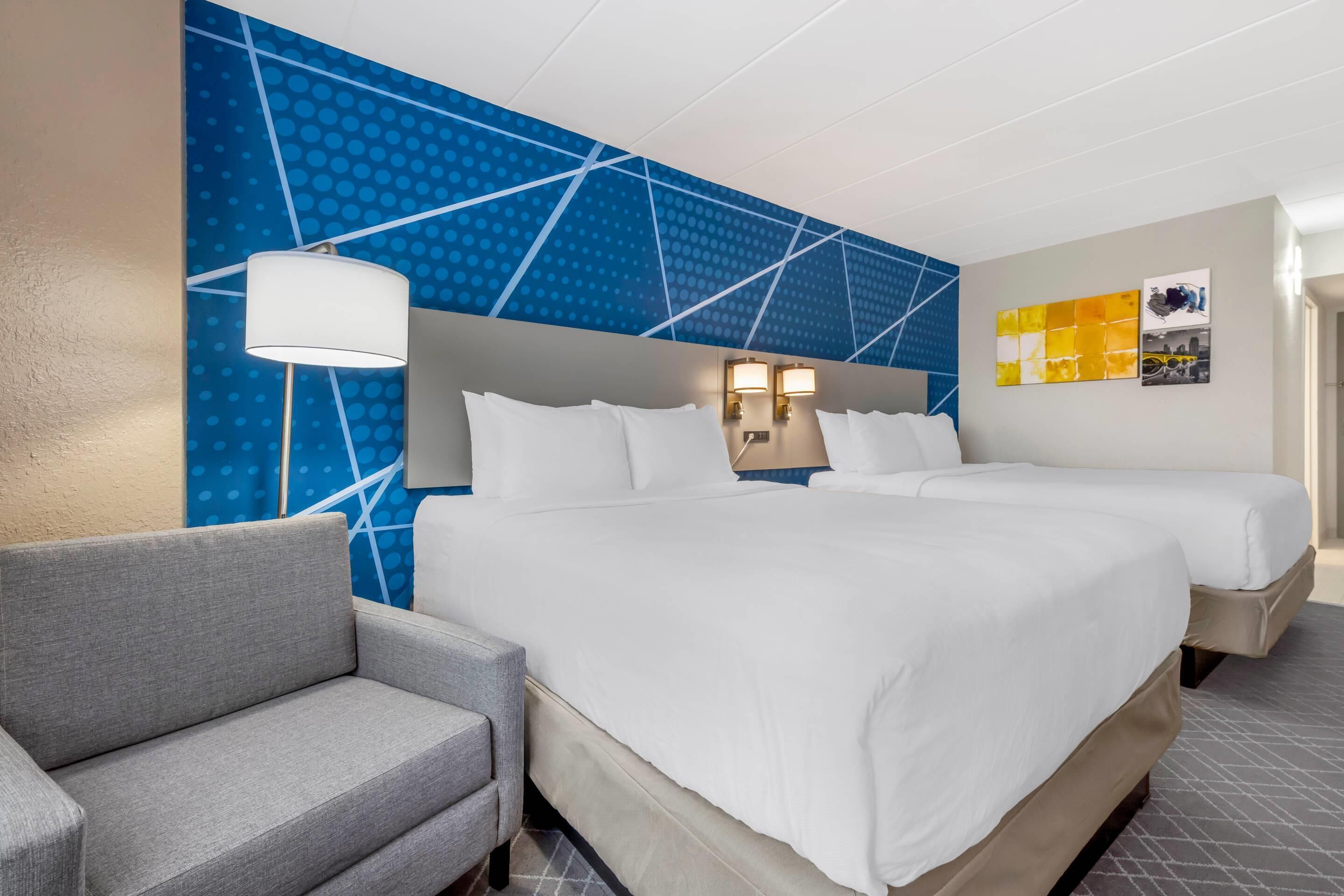 Une chambre d’hôtel avec deux lits, un canapé gris, un mur à motifs bleus et deux lampes fixées au mur.
