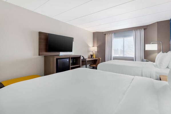 Ein Hotelzimmer mit zwei Betten, einem Fernseher, einem Schreibtisch mit Stuhl, einer Lampe und einem Fenster mit Vorhängen.