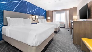 Ein Hotelzimmer mit zwei Betten, einem Schreibtisch, einem Fernseher und einem großen Fenster mit Vorhängen.