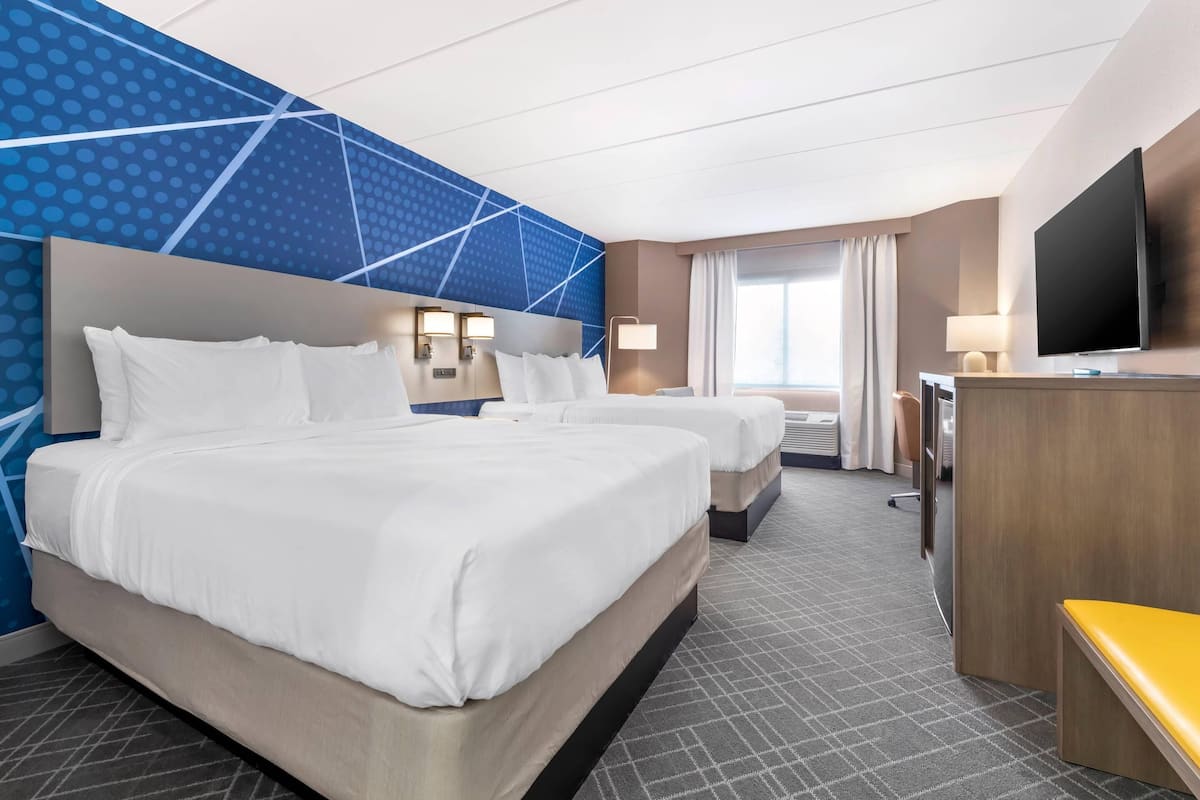A hotel room with two beds, a desk, a TV, and a large window with curtains.