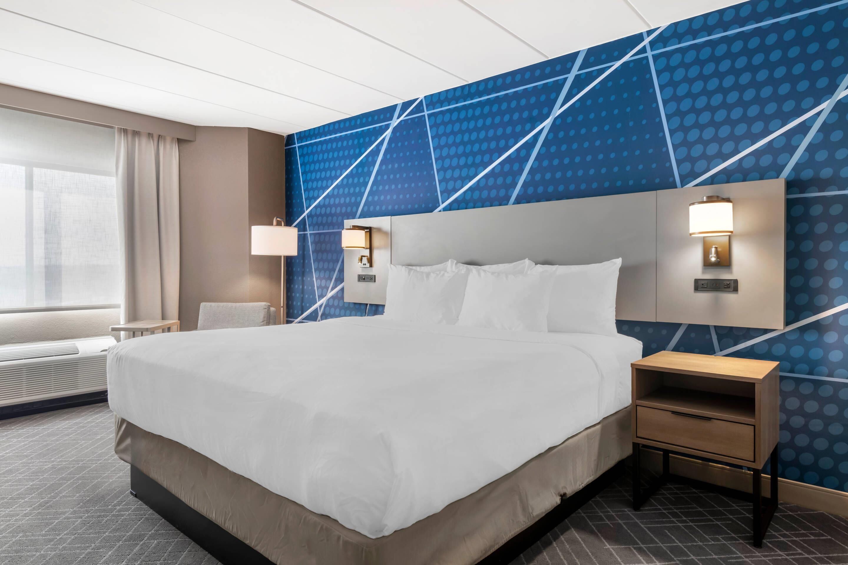 A hotel room with a large bed, bedside tables, a wall with a geometric pattern, and a window with curtains.