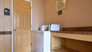 A laundry area with a washing machine, a dryer, and a countertop. There is a sign indicating a guest laundry room and a restroom sign on the wall. - Americas Best Value Inn Kingsville (Kingsville)