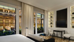 A hotel room with a large bed, a seating area with a table and chairs, a television, and a view of a building through large windows. - Hotel Bristol, a Luxury Collection Hotel, Vienna (Vienna)