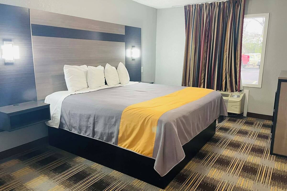 Una habitación de hotel moderna con una cama grande, una mesita de noche, cortinas en una ventana y una lámpara fijada a la pared.