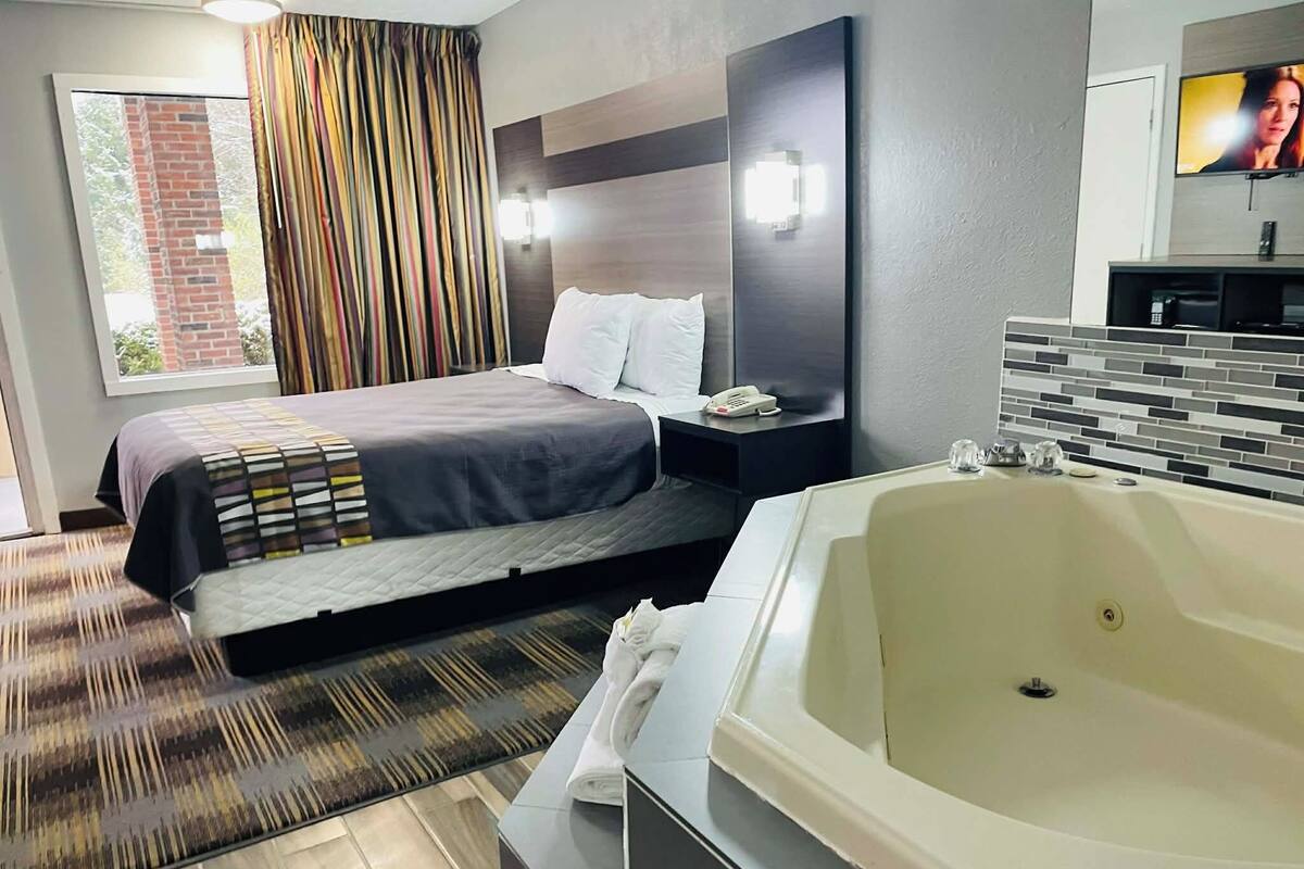 Una habitación de hotel moderna con una cama grande, un baño independiente con bañera, un televisor de pantalla plana y una ventana con cortinas.