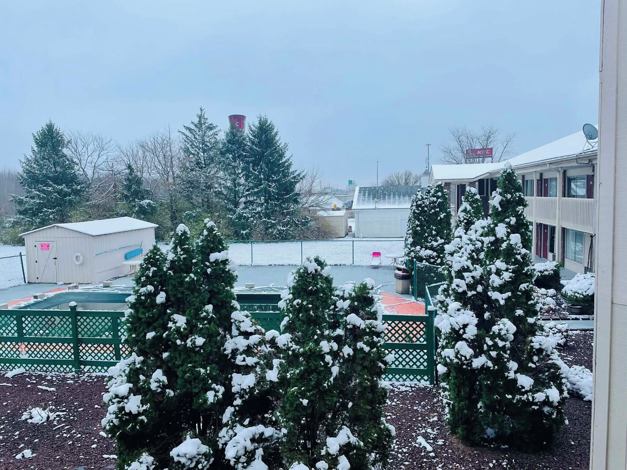 A snowy landscape with a building, trees, and a fenced area.