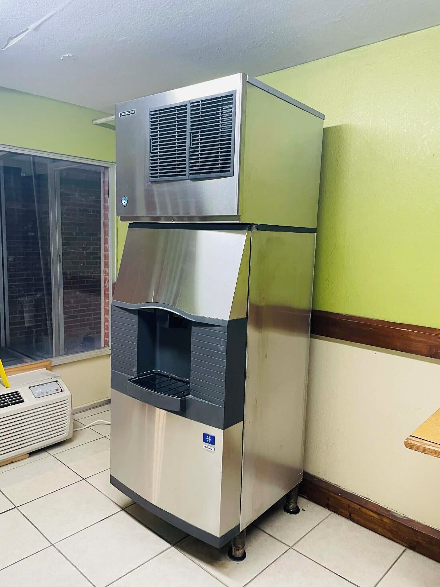 A commercial ice dispenser with a digital control panel, located in a room with green walls and a window.