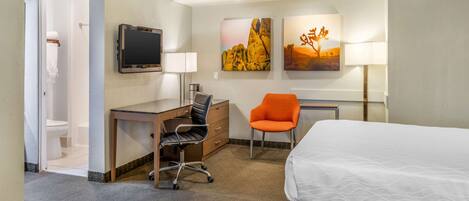 A hotel room with a bed, desk, chair, television, and artwork on the wall.
