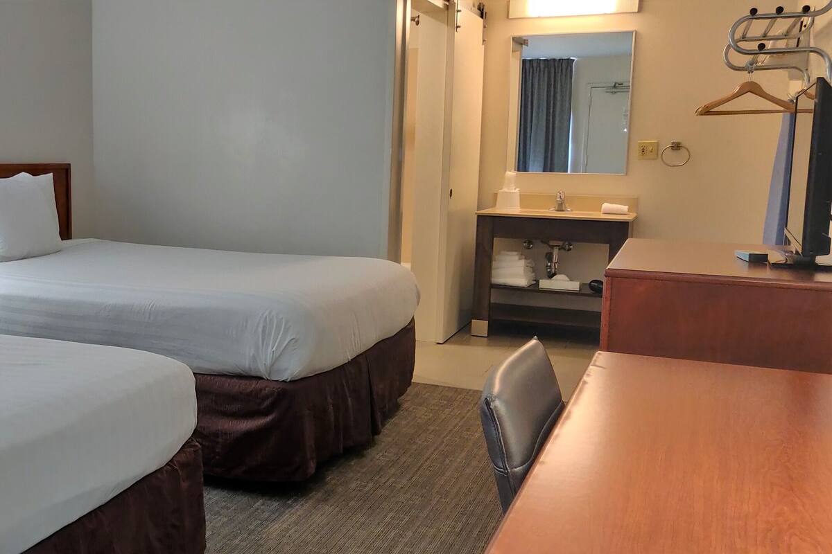 A hotel room with two single beds, a desk, a chair, a bathroom with a mirror, and a coat rack.