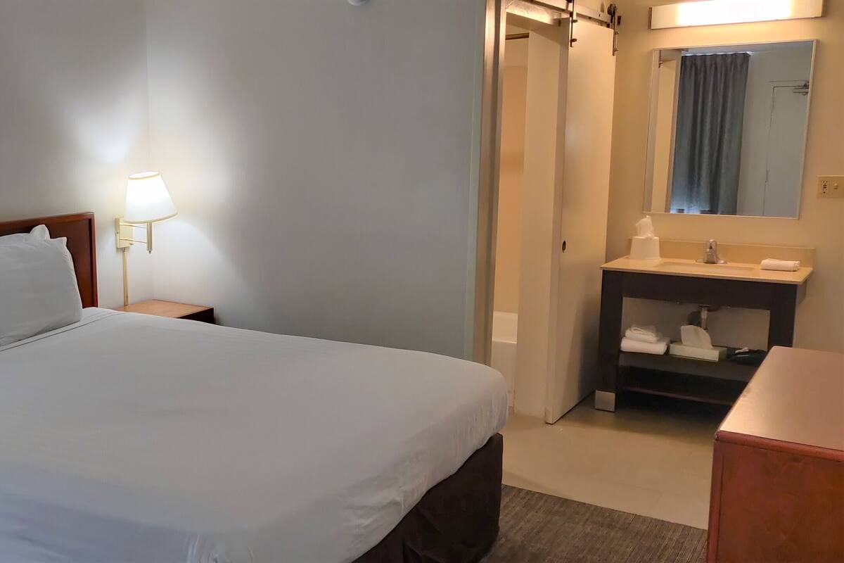 A hotel room with a bed, two bedside lamps, a nightstand, a bathroom with a sink and mirror, and a wooden dresser.