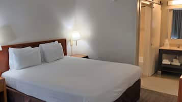 Una habitación de hotel con una cama, dos lámparas de noche, una mesita de noche, un baño con lavamanos y espejo, y un aparador de madera.