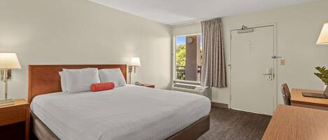 A hotel room with a large bed, a desk with a laptop, a window with curtains, and a door.