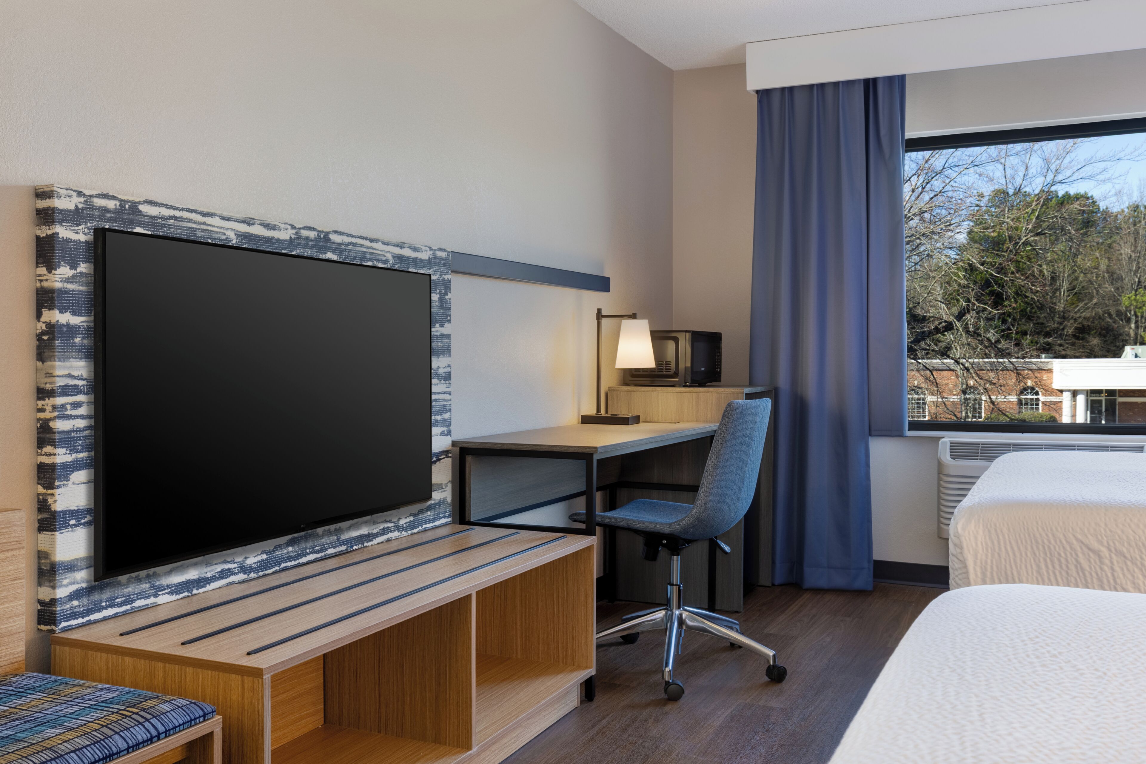 A hotel room with a bed, a desk with a chair, a television, and a window with curtains.