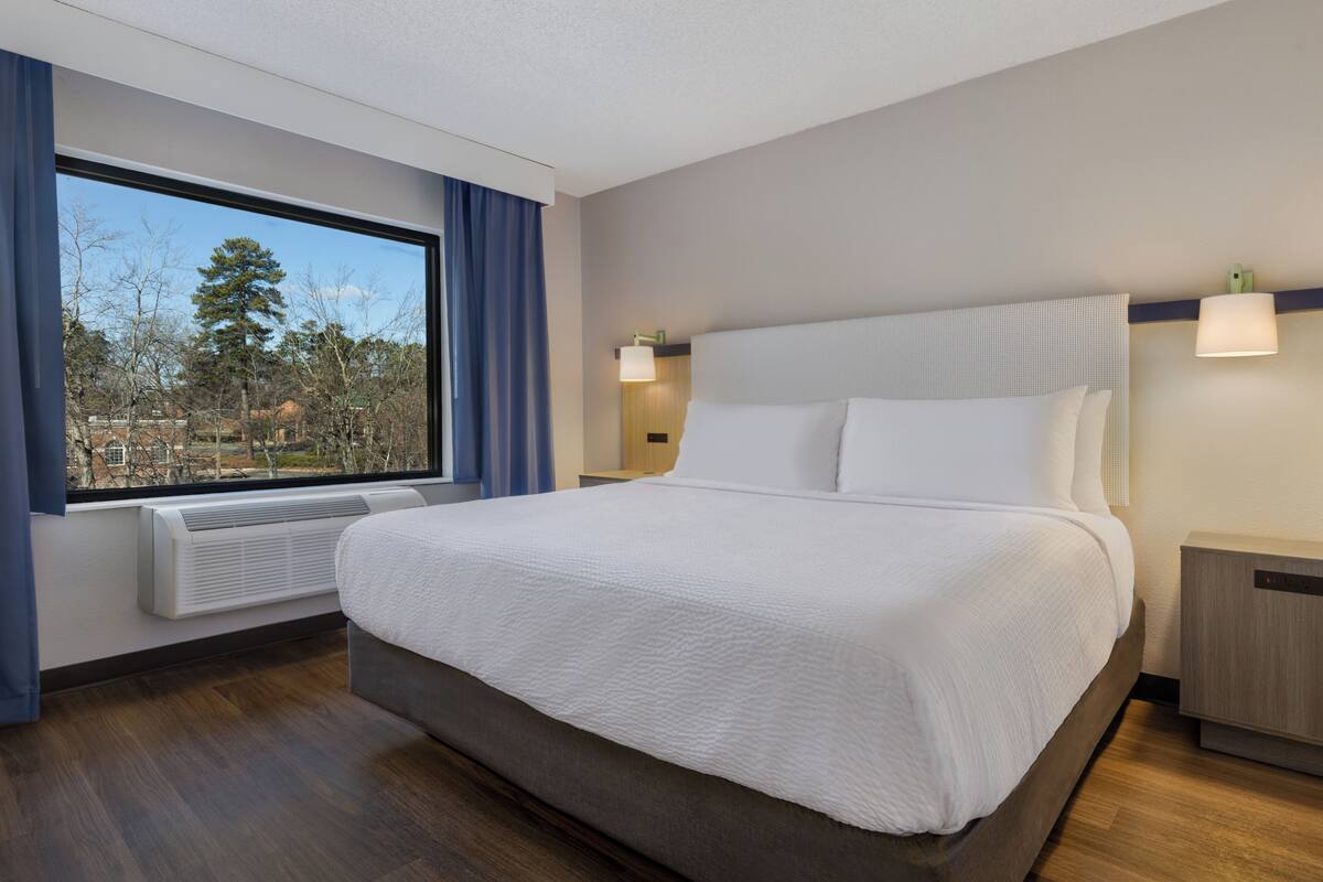 A hotel room with a large bed, a window with curtains, a wall-mounted heater, and a bedside table.