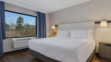 A hotel room with a large bed, a window with curtains, a wall-mounted heater, and a bedside table.