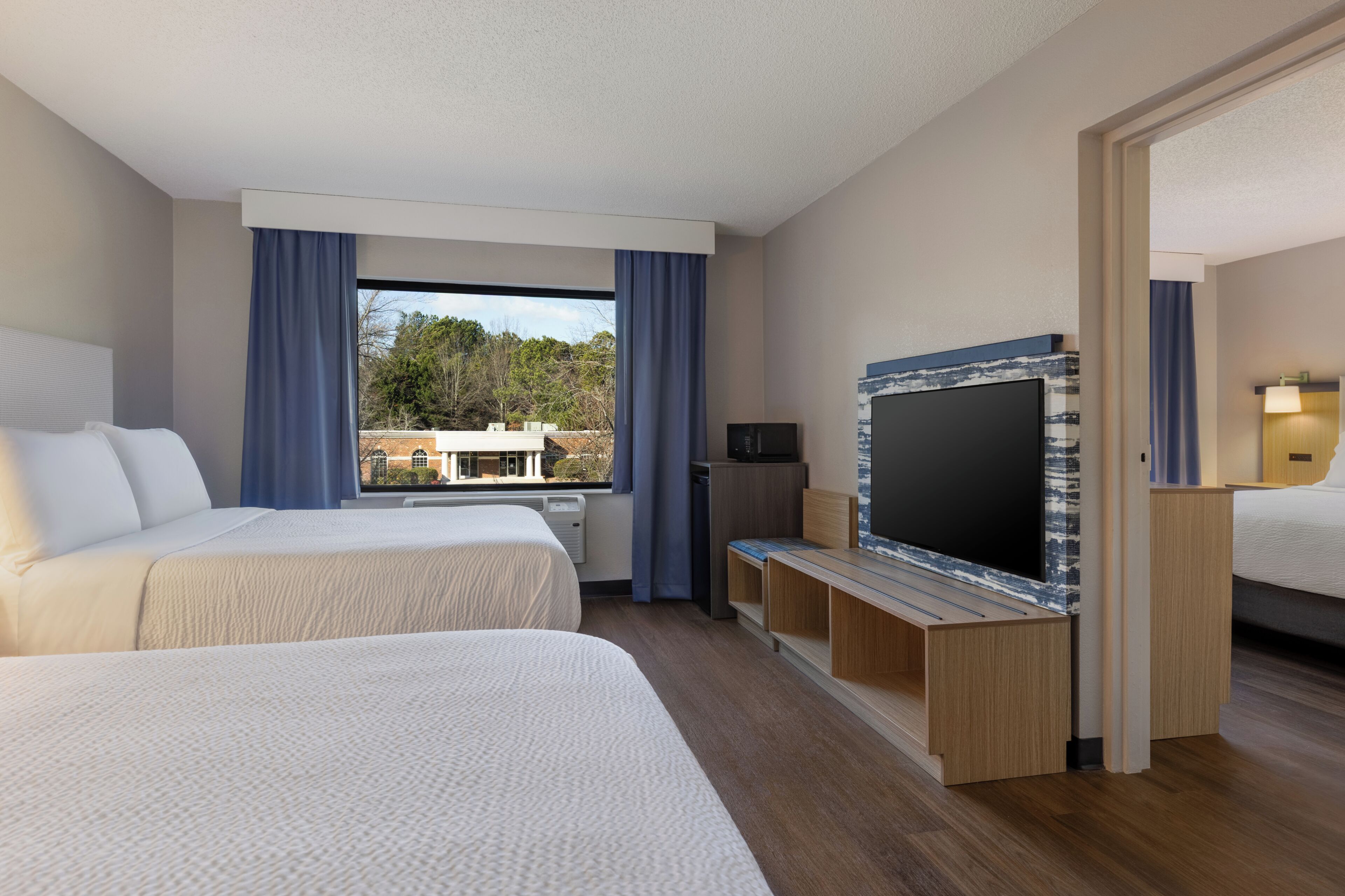 Hotellihuone, jossa on kaksi sänkyä, televisio ja näkymä ulkopuolella olevaan rakennukseen ja puihin.