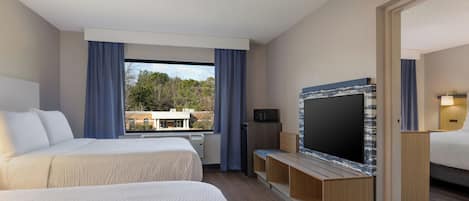 Hotellihuone, jossa on kaksi sänkyä, televisio ja näkymä ulkopuolella olevaan rakennukseen ja puihin.