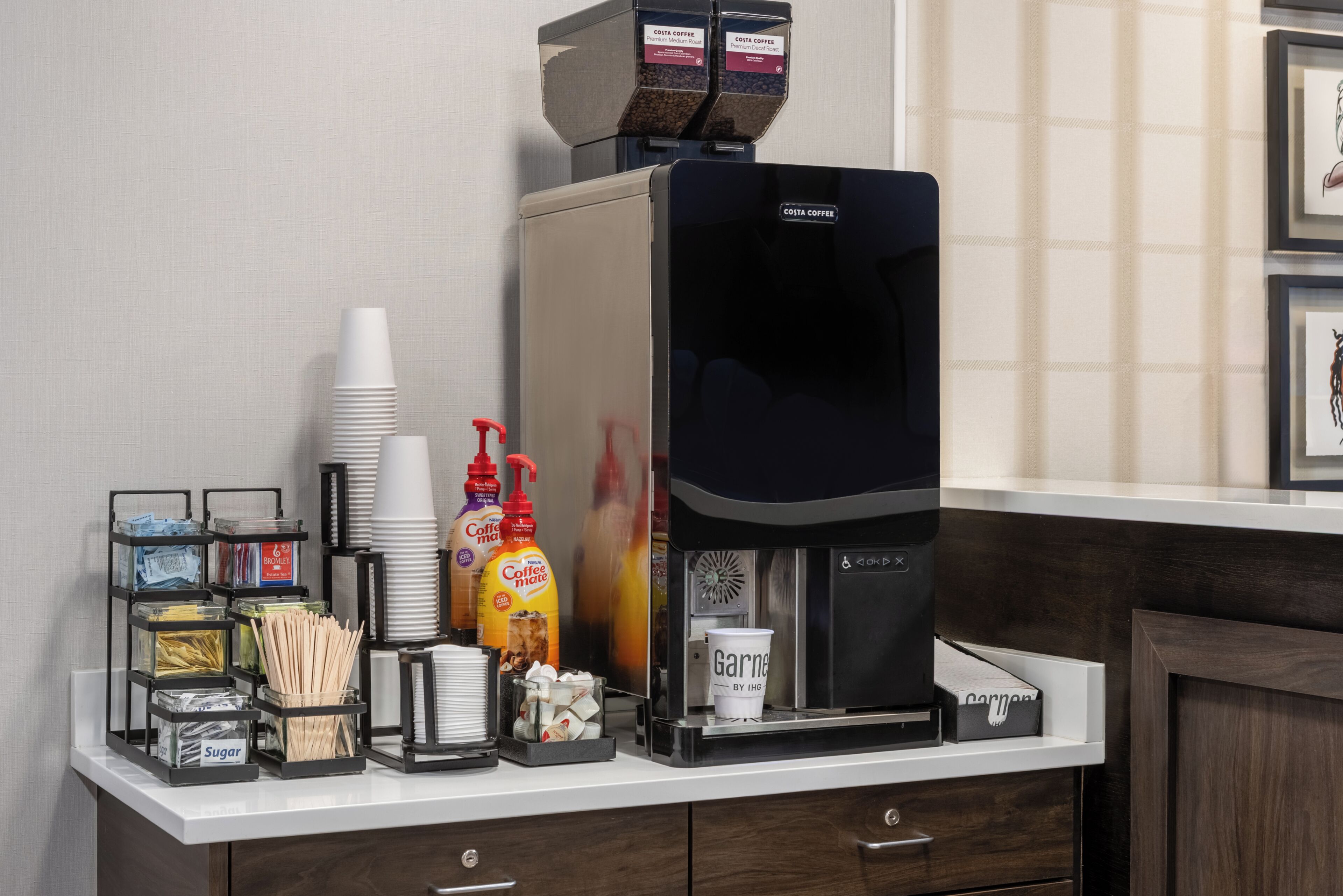 A coffee station with a coffee machine, disposable cups, sugar, coffee creamer, and a coffee grinder.