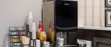 A coffee station with a coffee machine, disposable cups, sugar, coffee creamer, and a coffee grinder.