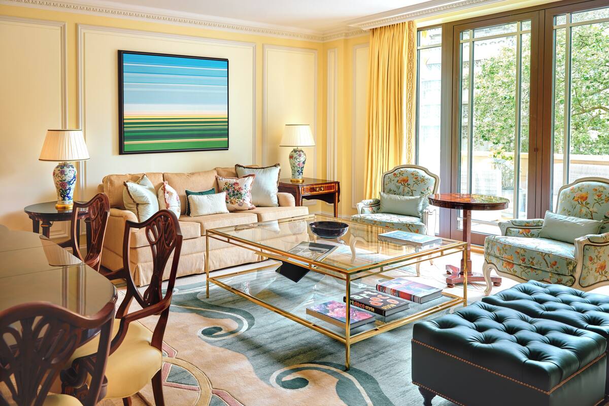 A well-lit living room with a glass coffee table, leather sofa, floral armchairs, and a view of greenery outside.