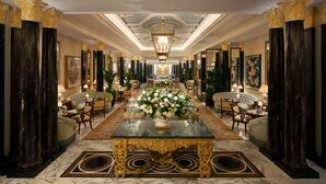 Un lobby de hotel elegante con una mesa de vidrio en el centro, columnas ornamentadas y un candelabro.