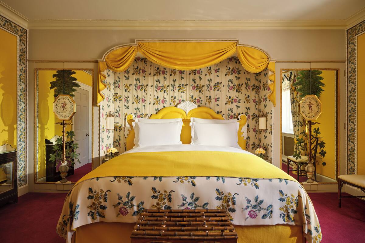 Una cama grande con cabecera floral y cortinas amarillas, un banco de madera y un espejo con marco floral.